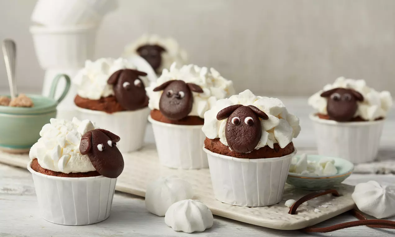 Cupcakes en caissettes blanches avec des visages de moutons en chocolat brun surmontés de crème blanche aérienne, disposés sur un plateau