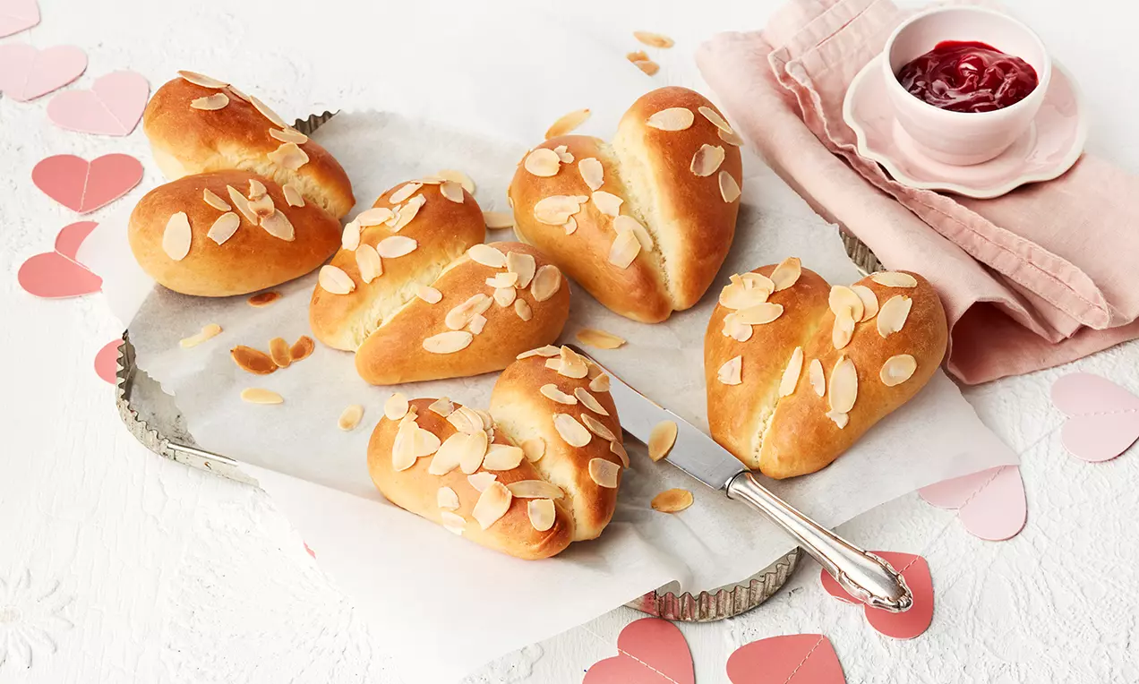 Goldgelbe, herzförmige Hefe-Brötchen mit glänzender Oberfläche und gerösteten Mandelblättchen auf Backpapier mit weißer Stoffserviette