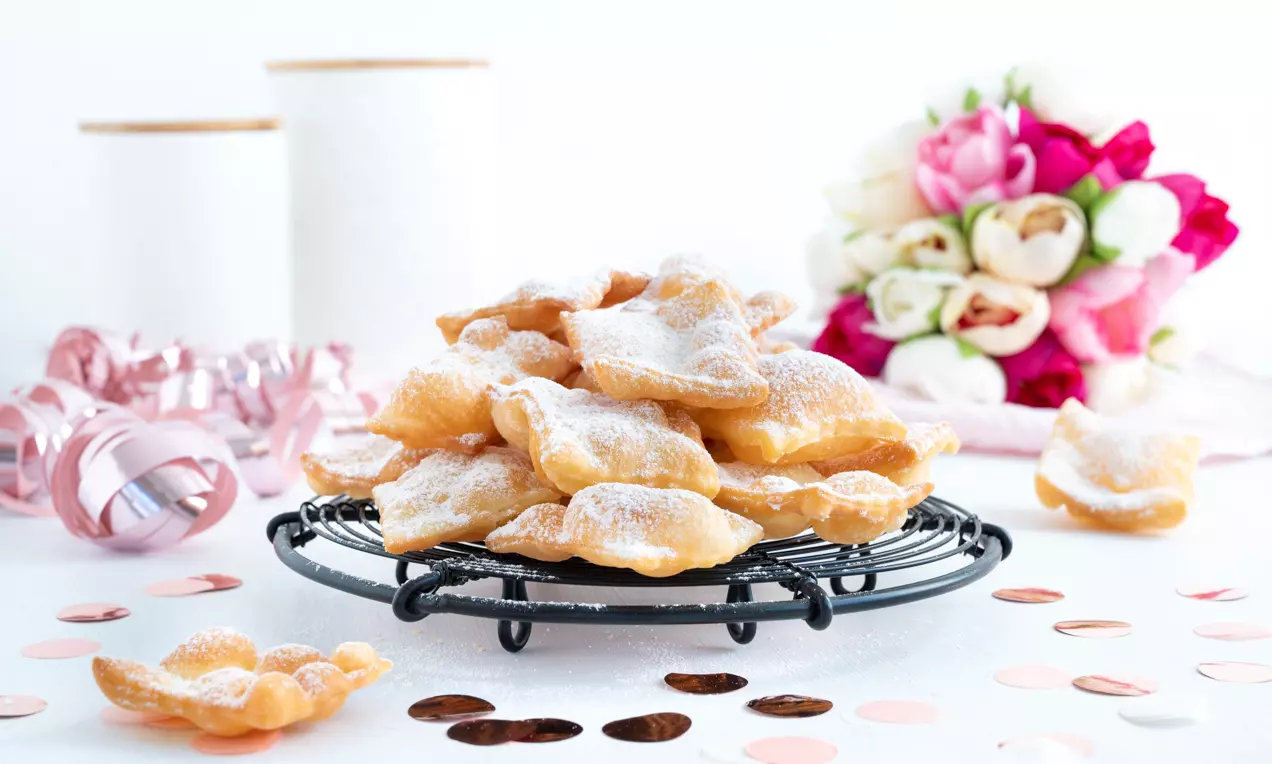 Goldbraune, knusprig-dünne Fasnachts-Chüechli unregelmäßiger Form, mit Puderzucker bestäubt auf schwarzem Gitter, umgeben von roséfarbener Dekoration