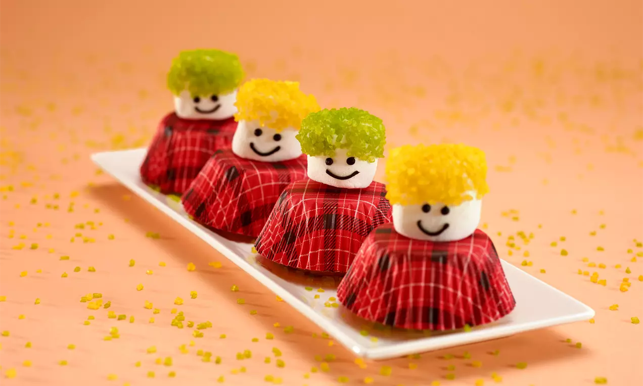Muffins vêtus de caissettes écossaises rouges, coiffés de marshmallows souriants et de bonbons éclatants jaunes et verts sur un plateau blanc