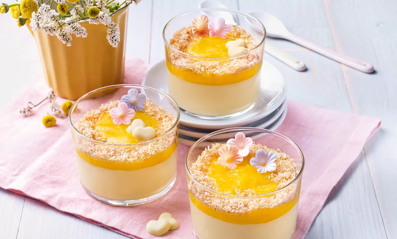 Kokos-Pudding in Gläsern mit leuchtend gelbem Mangopüree, gerösteten Kokosraspeln und bunten Blüten aus Marzipan auf rosa Stoff drapiert