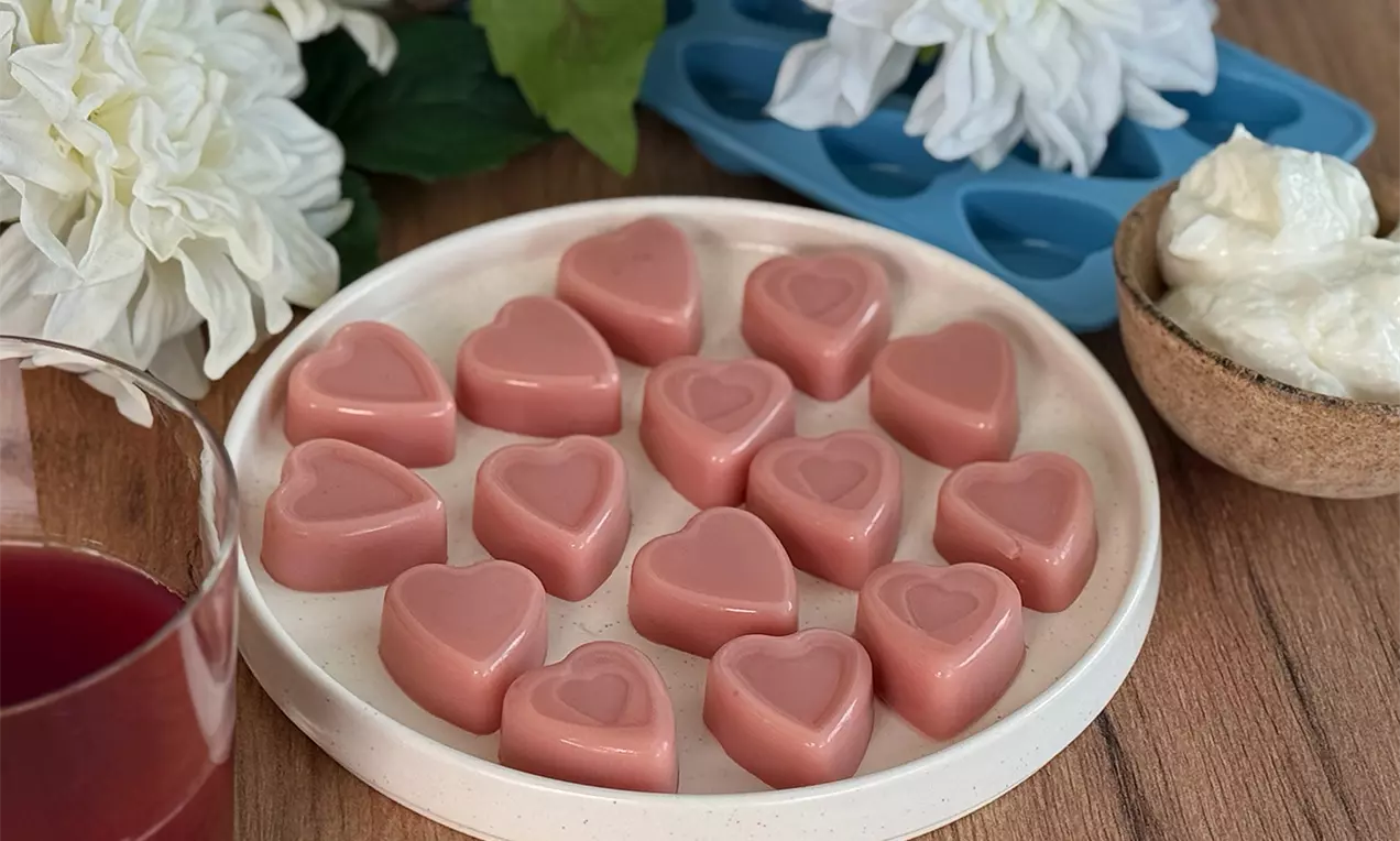 Fruchtige rosa Gummi-Herzen, die auf einem beigen Teller auf einem Holztisch liegen. Dekortiert mit einer weissen Blume, einem Glas rotem Saft und einer Schale mit Joghurt