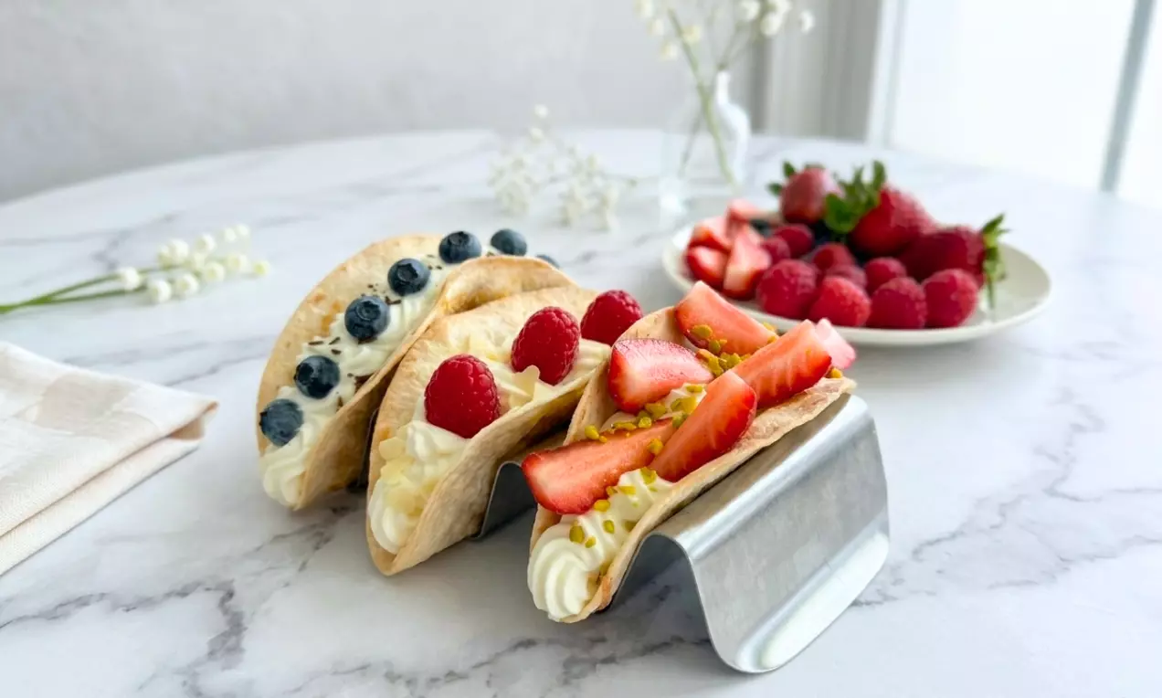 Süße Tacos mit goldbrauner Teighülle, gefüllt mit weißer Sahne, dekoriert mit Beeren und Pistazien auf silbernem Halter auf Marmortisch