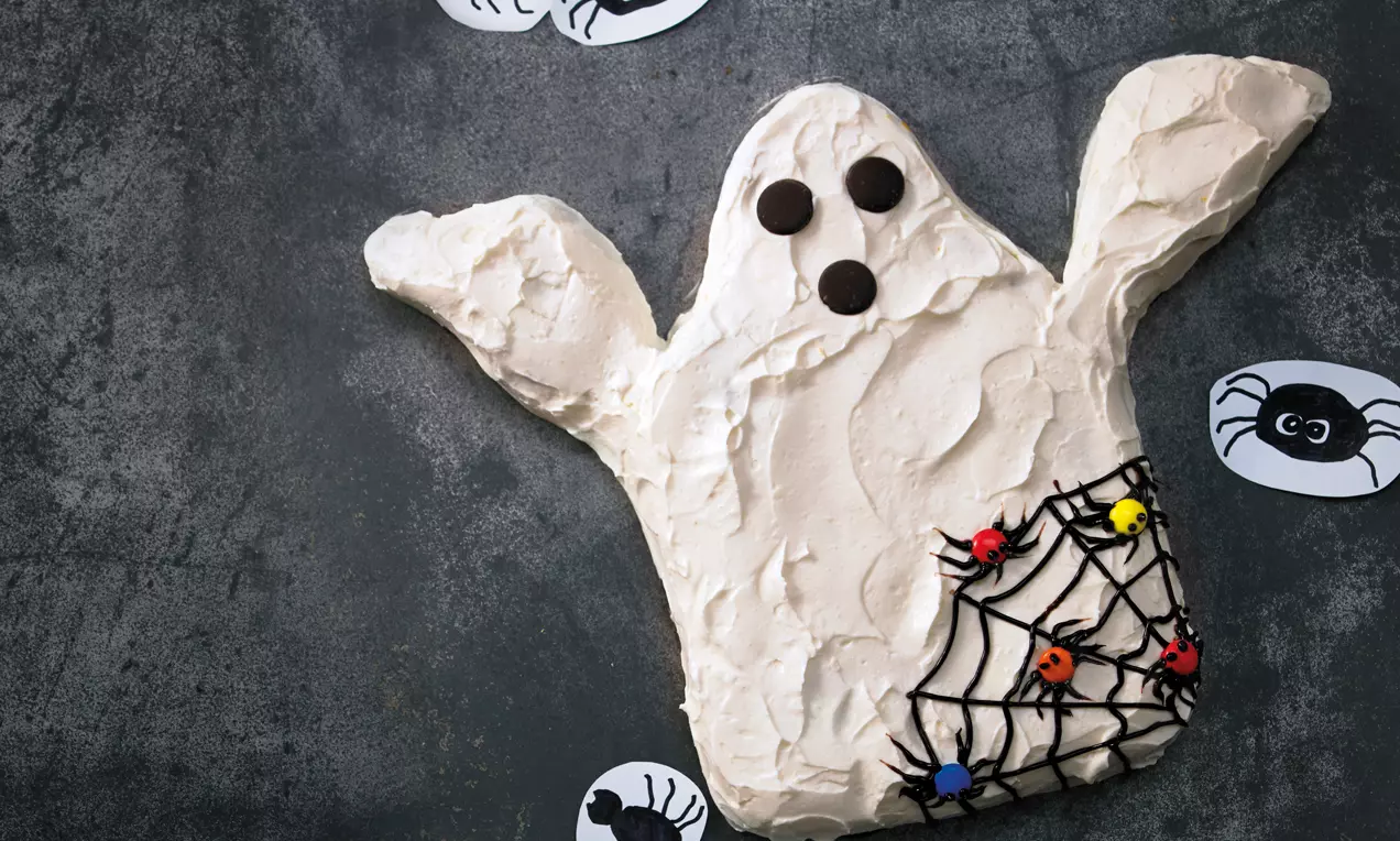 Gâteau en forme de fantôme blanc texturé avec toile d'araignée chocolatée, boules colorées et yeux en pastilles sur une table sombre