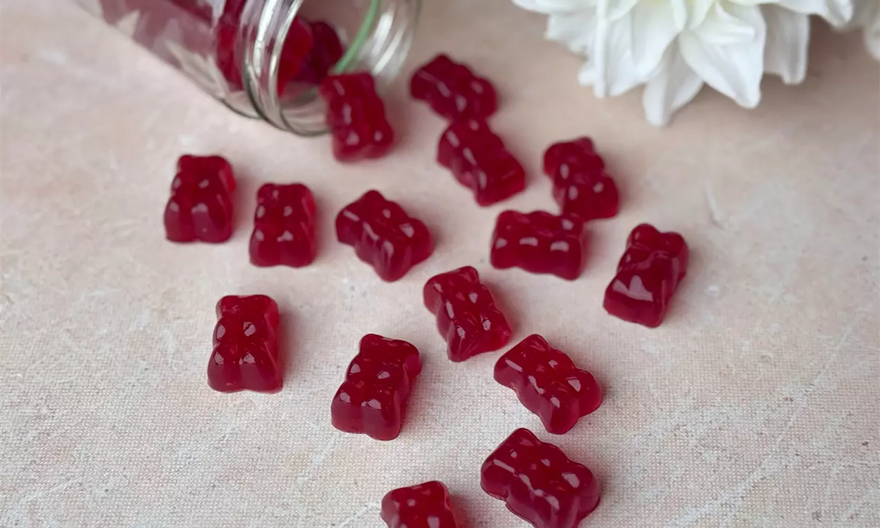 Rote, glänzende Gummibärchen in Bärenform verstreut auf hellrosem Untergrund neben einem umgekippten Glas und weißen Blüten