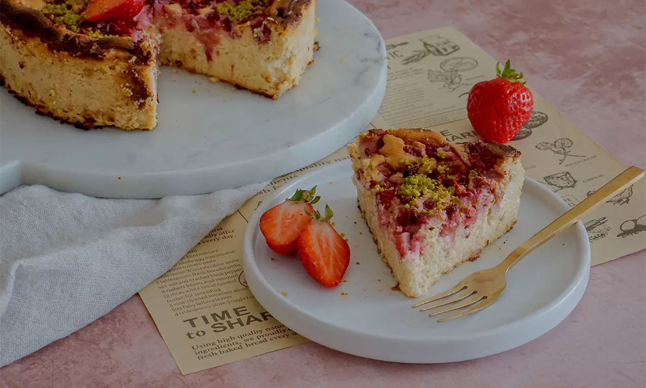 Ein Stück Protein-Cheesecake mit goldbrauner Kruste, rosafarbenem Fruchtbelag, Pistazien und Erdbeerscheiben auf einem weißen Teller mit goldener Gabel