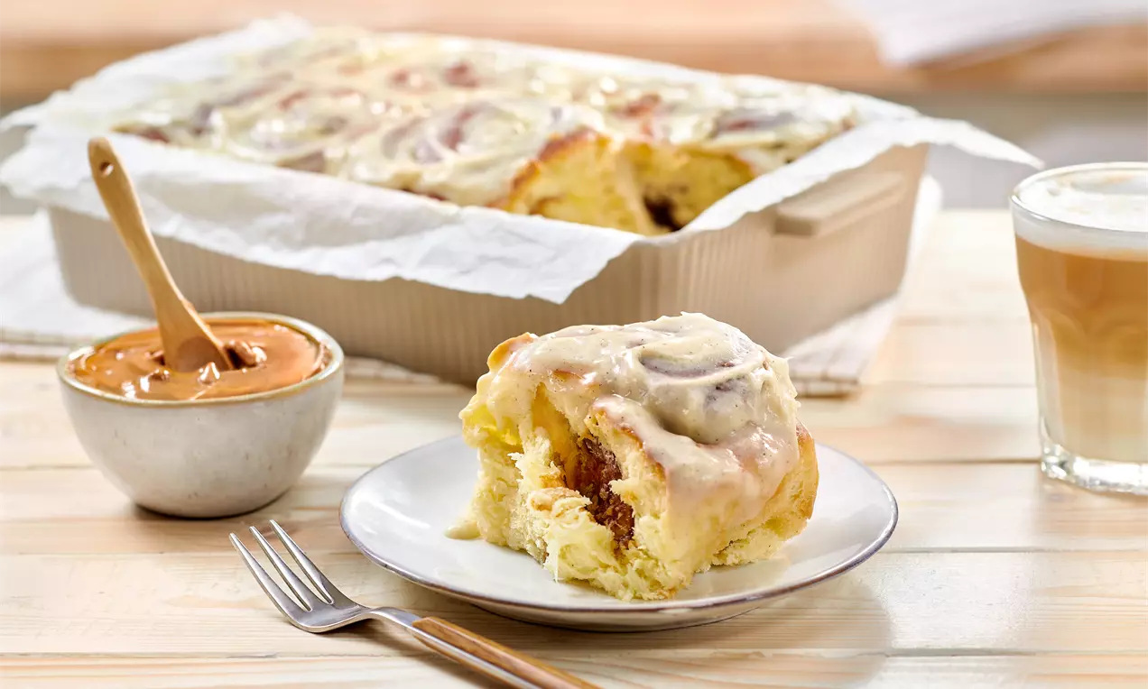 Roulés dorés à la cannelle glacés légèrement brillants sur une assiette blanche avec crème caramel et café latte en arrière-plan