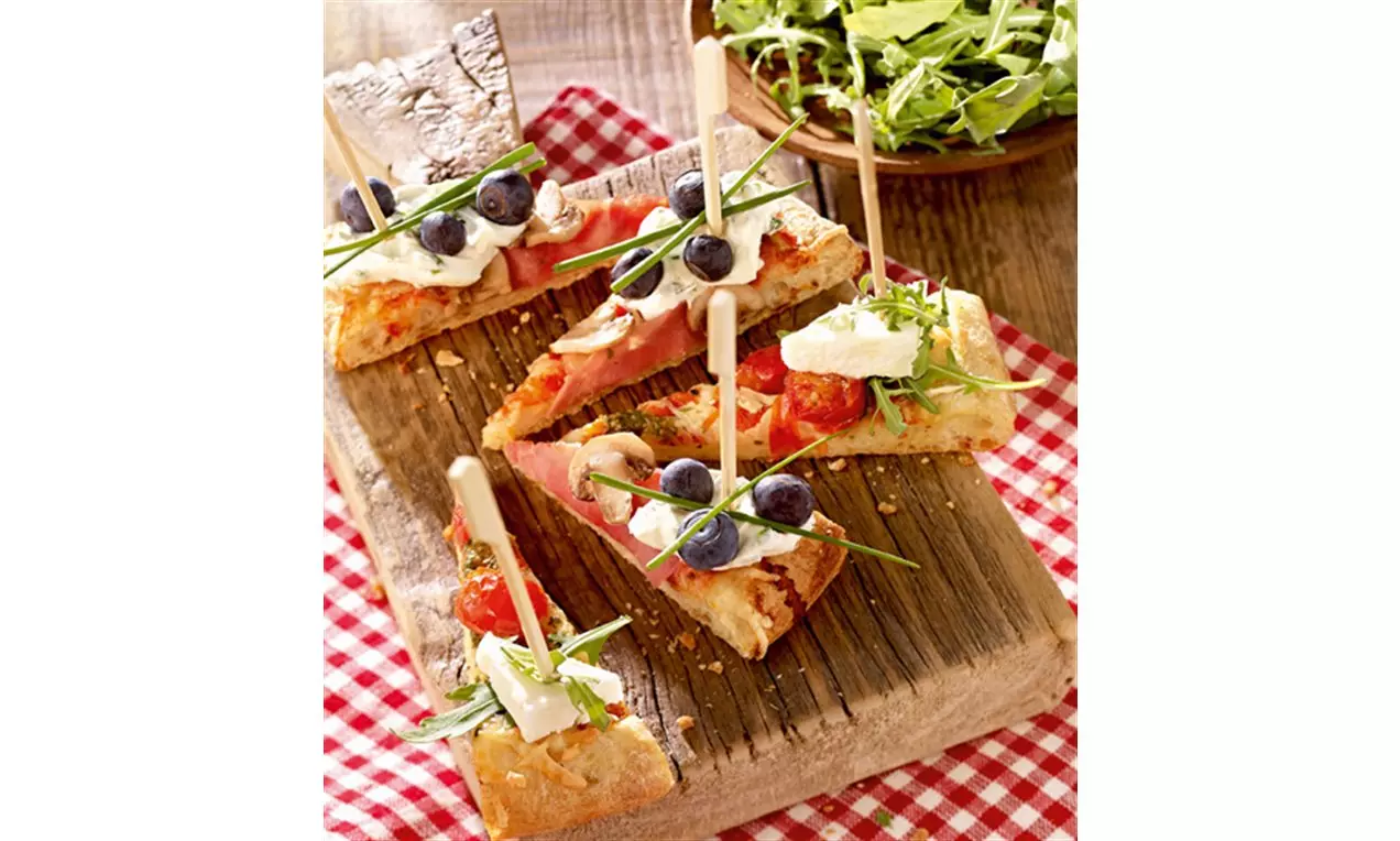 Goldbraune Mini-Pizzastücke auf Holzbrett, mit Spießen fixiert, belegt mit Salat, Tomaten, Schinken, Blaubeeren und frischen Kräutern