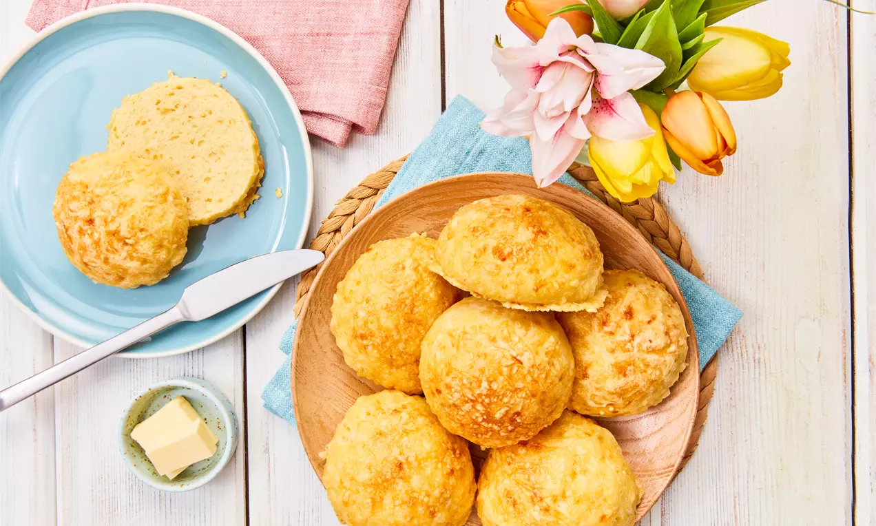 Goldbraune, runde Käsebrötchen auf einem Holzteller, begleitet von einer rosa Serviette, Butterstücken und bunten Blumen