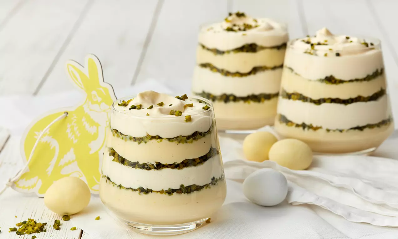 Schichtdessert im Glas mit hellgelbem Eierlikör-Pudding, weißer Creme, gehackten Pistazien und Dekoration aus Osterfiguren und bunten Eiern