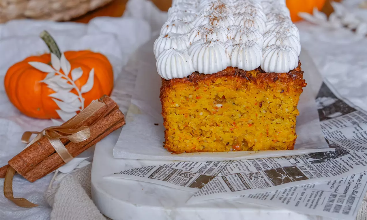 Kürbis-Haselnuss-Cake mit saftig goldgelbem Inneren, dekoriert mit weißen Sahnetupfen und Zimt auf Marmorplatte mit herbstlicher Dekoration