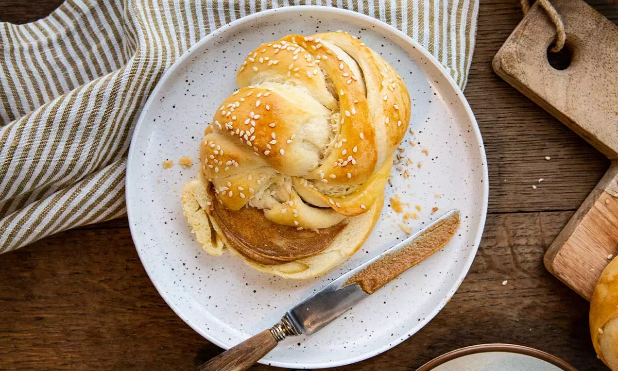 Pane turco intrecciato dorato con semi bianchi, dalla trama morbida e soffice, su un piatto bianco accanto a un coltello di legno rustic