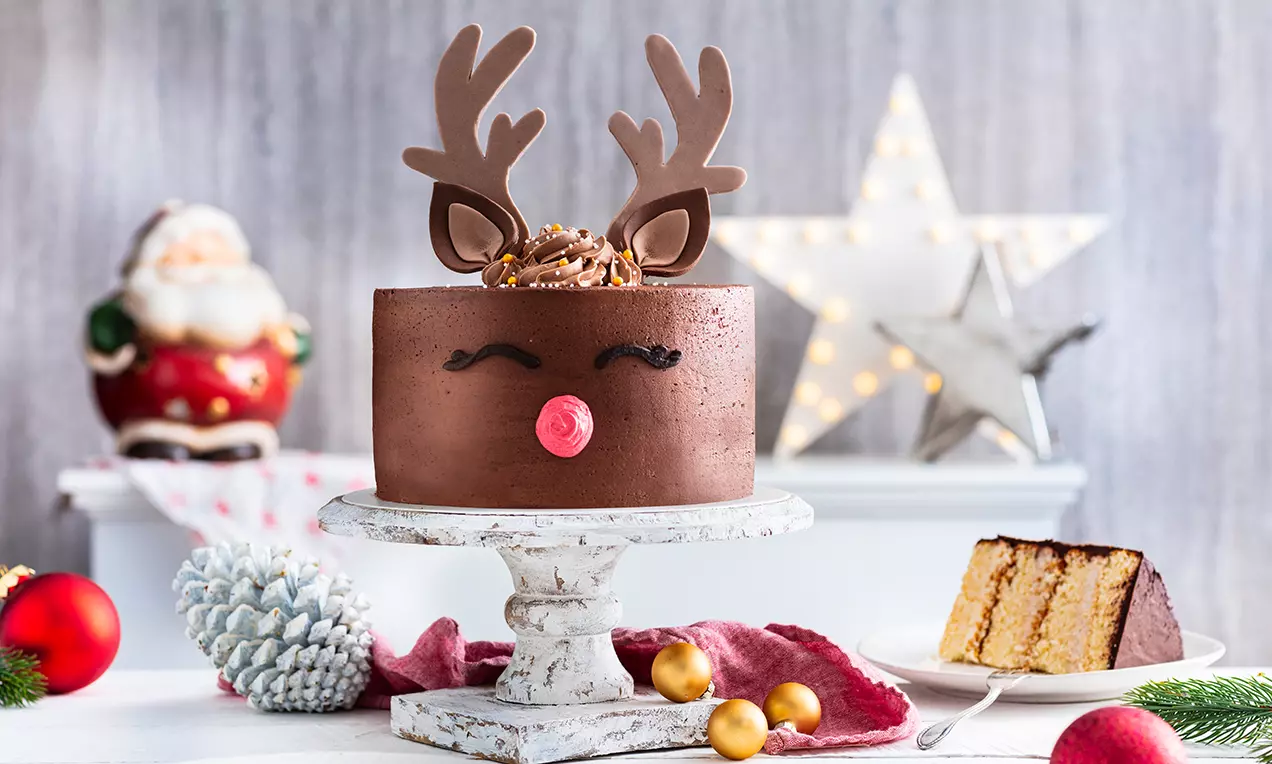 Runde Rentier-Torte aus brauner Schokoladenglasur mit roten Zuckernase, schokoladigen Geweihen und filigranen Verzierungen auf rustikalem Tortenständer