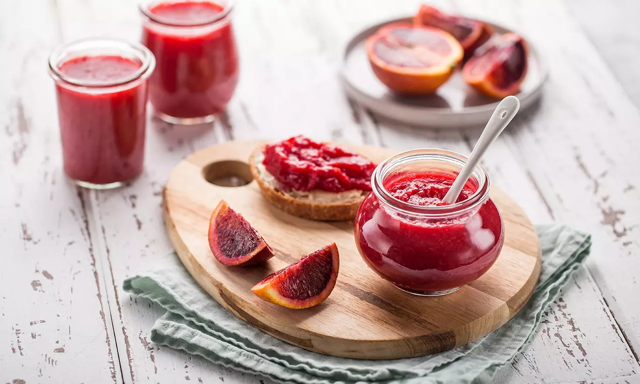 Blutorangen Konfitüre in einem runden Glas mit weißem Löffel auf Holzbrett, daneben frische Blutorangen und bestrichene Scheibe auf hellgrünem Tuch