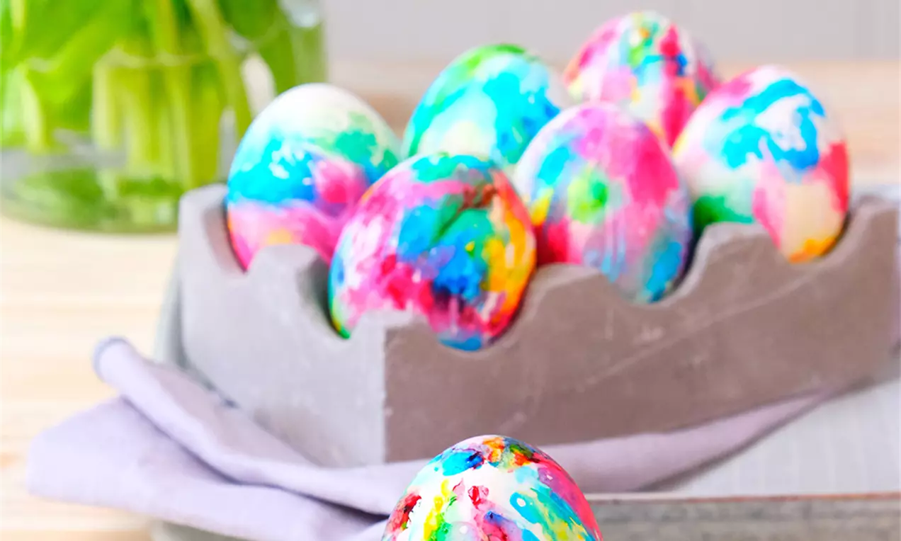 Bunte Batik-Ostereier mit lebhaften Farbmustern in einer grauen Eierbox, ein einzelnes Ei dekorativ in einer weißen Porzellanschale