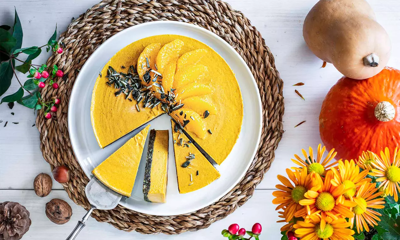 Cheesecake orange vif sans cuisson, garni de graines foncées et de fines tranches de courge sur une assiette blanche entourée de décorations automnales