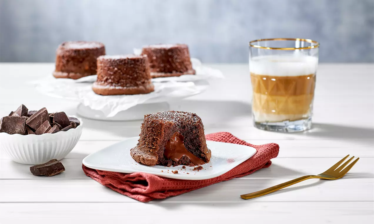 Schokoküchlein mit flüssigem Kern auf weißem Teller, daneben goldene Gabel, rotes Stofftuch, dunkle Schokoladenstücke und Latte macchiato