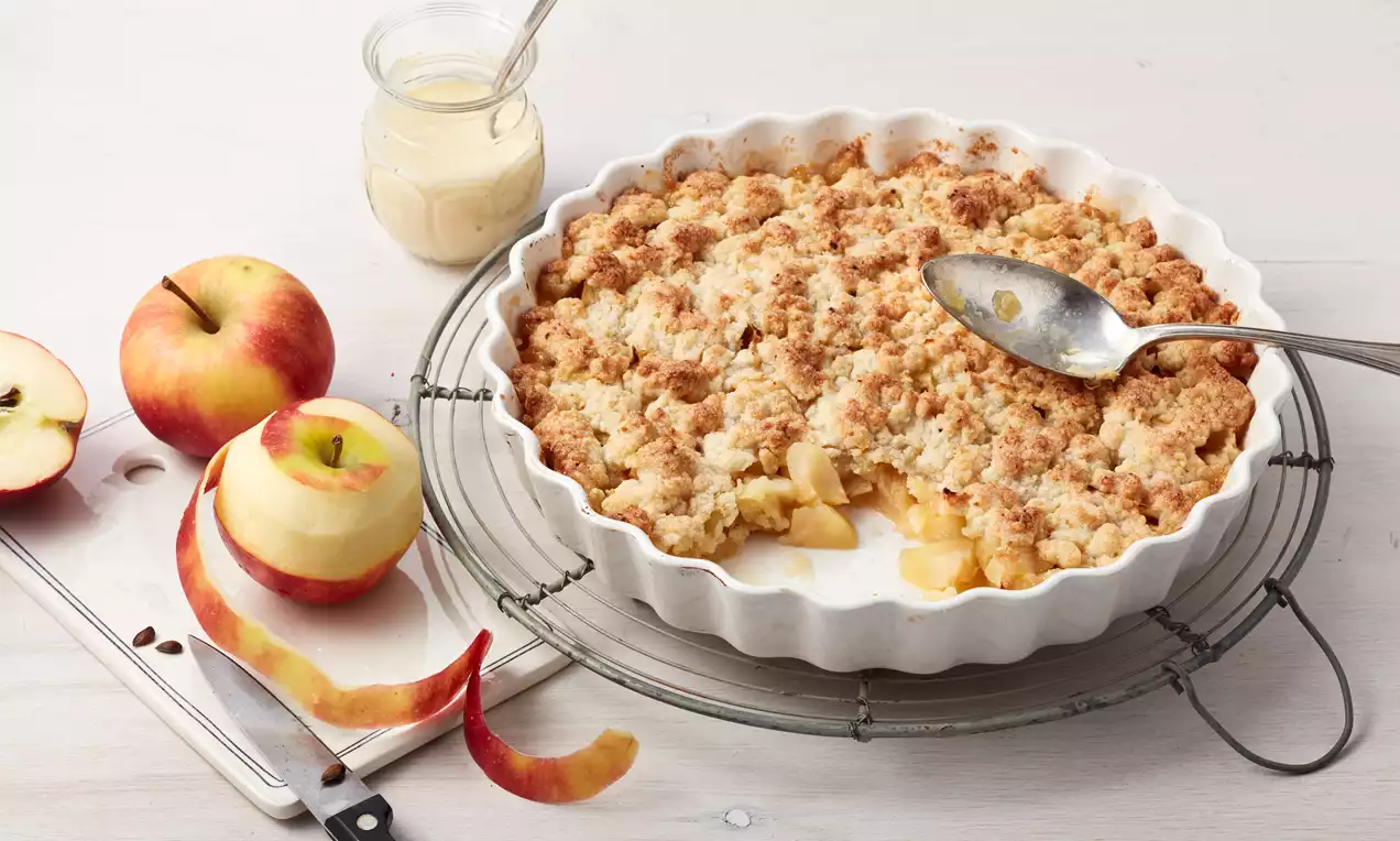 Goldbraune Streusel mit zarten Apfelstücken in einer weißen runden Form, daneben frische Äpfel mit Schale und ein Glas Vanillesauce