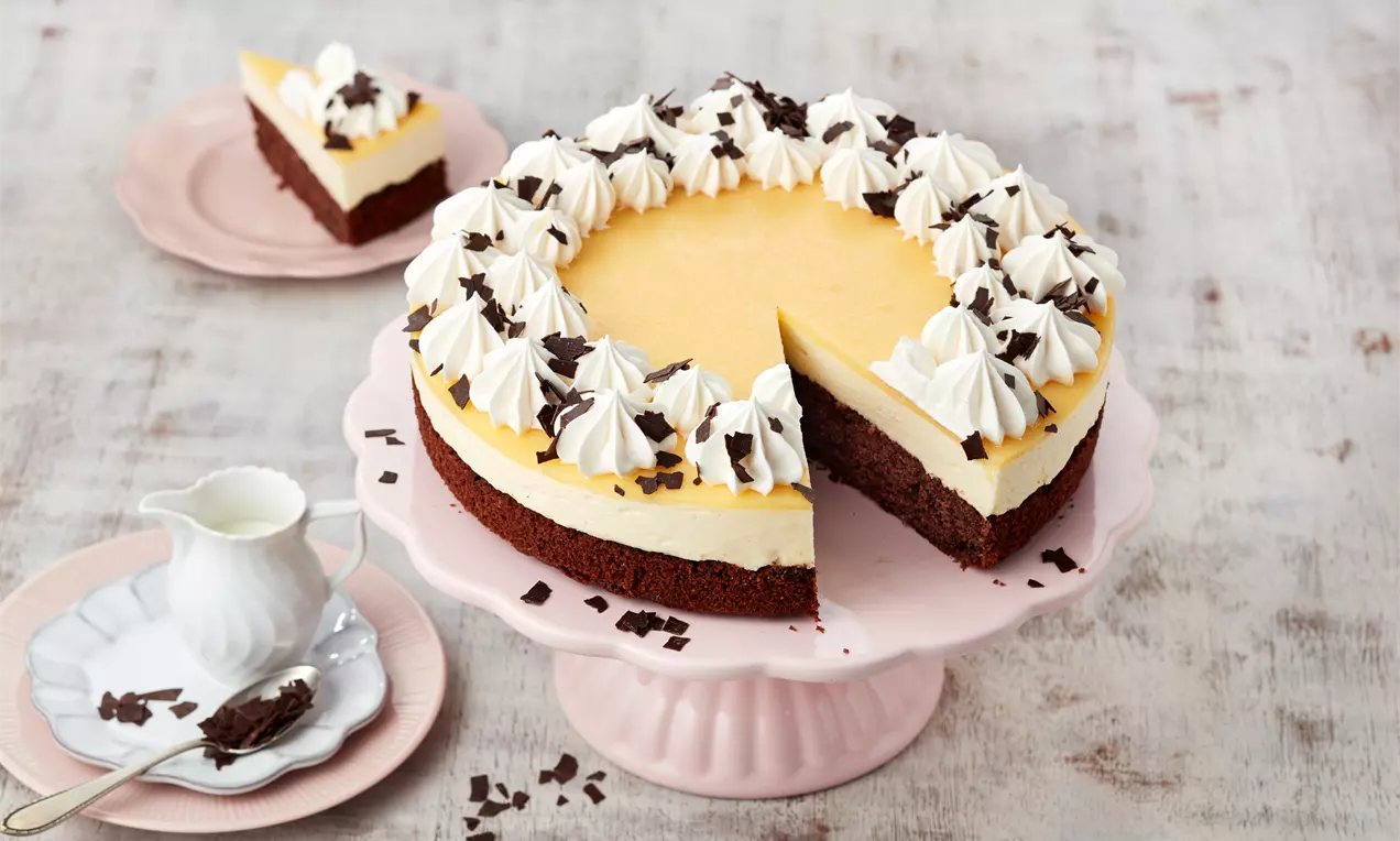 Eierlikörtorte mit gelber Creme, sahnigen Tupfen und Schokoraspeln auf dunklem Boden, serviert auf rosa Tortenplatte mit Teller und Kännchen