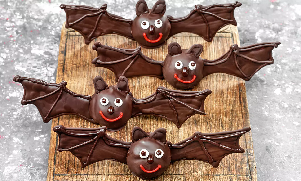 Chauves-souris en chocolat noir brillant avec ailes étendues et petits visages souriants, posées sur une planche en bois rustique