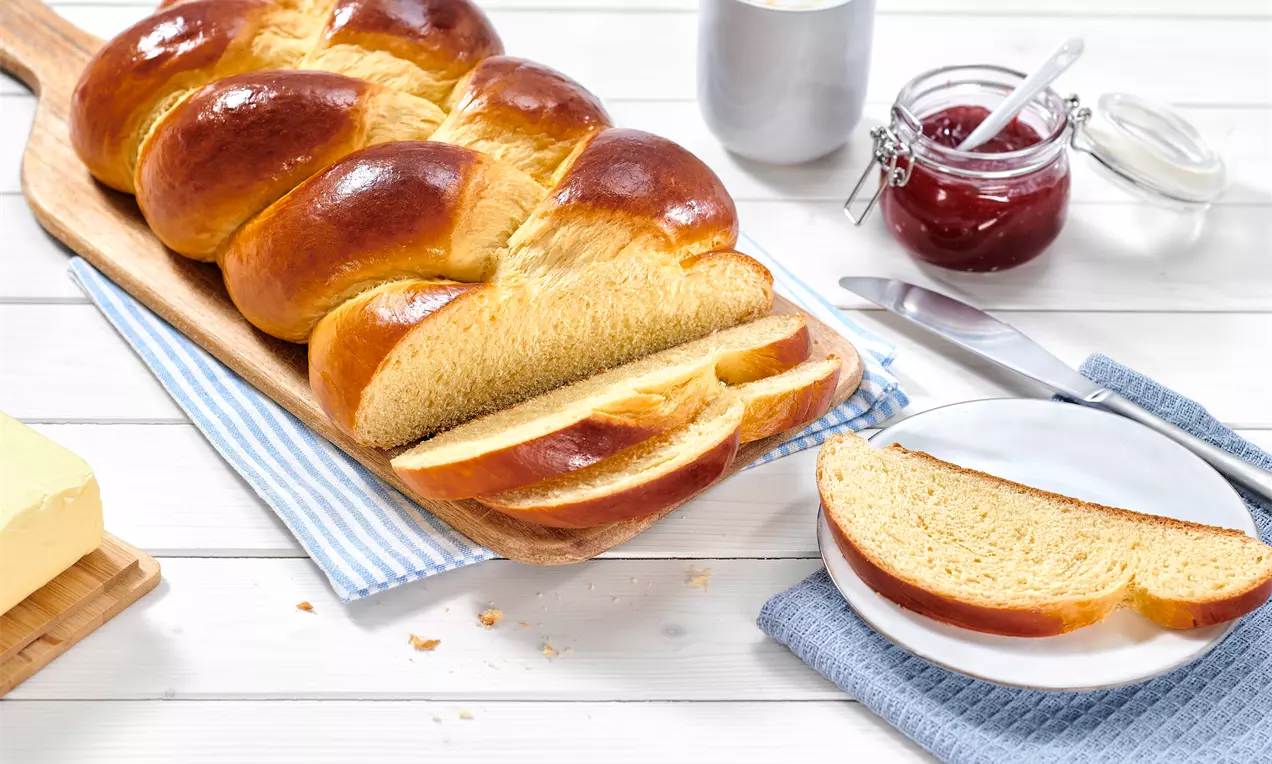 Hefezopf mit glänzender, goldbrauner Kruste und weicher Textur auf einem Holzbrett, angeschnitten neben Butter, Marmelade und Sahne