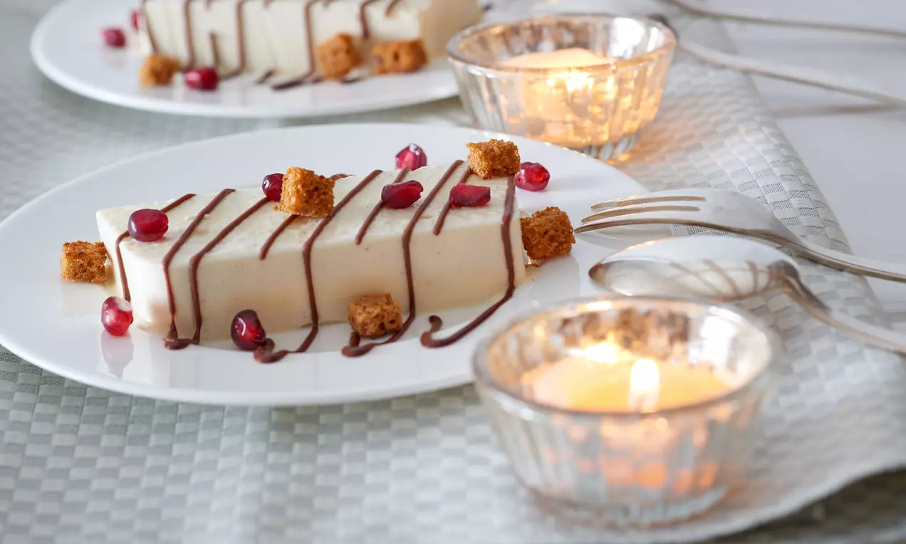 Rechteckiges Lebkuchen-Parfait mit glatter Oberfläche, Schokoladenstreifen, granatrote Granatapfelkerne und braune Kekswürfel auf weißem Teller