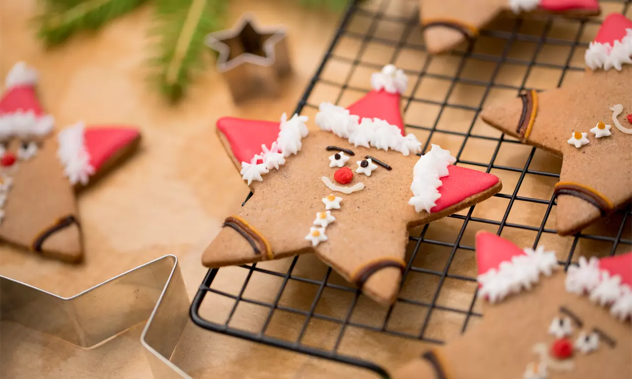 Lebkuchen Chläuse aus braunem Teig mit roten Zuckermützen, weißer Zuckerdekoration und kleinen Gesichtsförmchen auf einem schwarzen Gitter