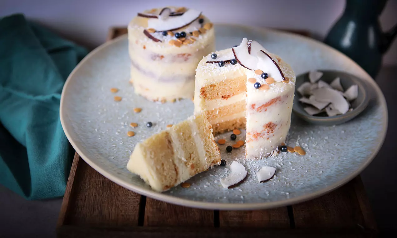 Mini gâteaux à étages à la crème blanche, ornés de perles argentées et copeaux de noix de coco sur une assiette beige saupoudrée de sucre