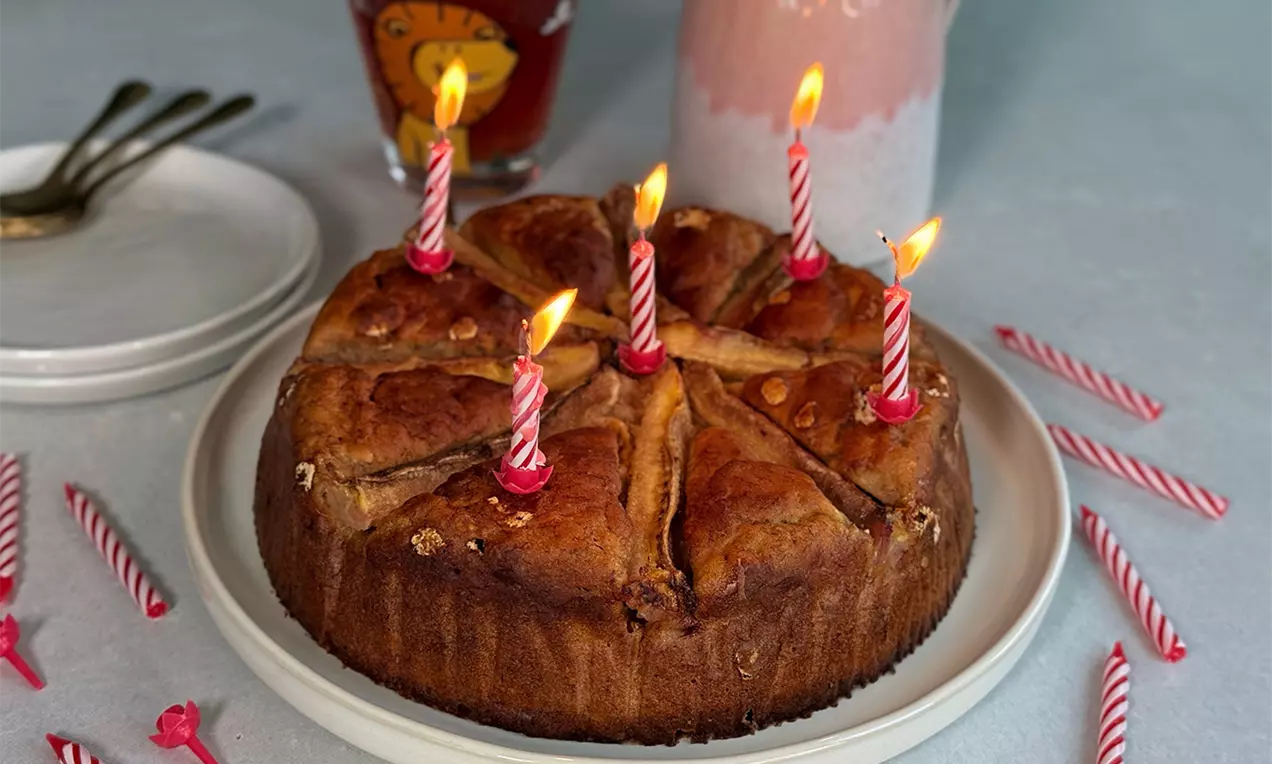 Goldbrauner Bananenkuchen mit gebrannten Kerzen auf weißem Teller, umgeben von roten Geburtstagskerzen und Geschirr auf hellblauer Tischplatte