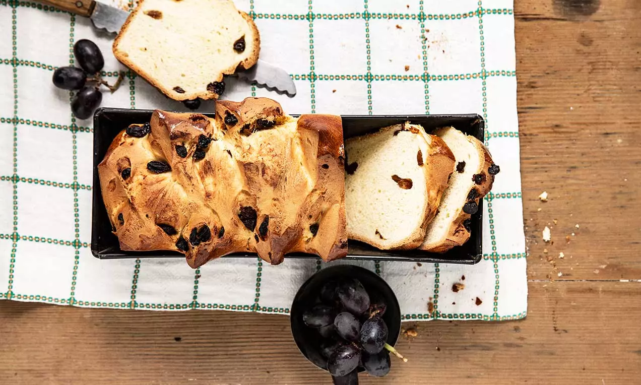 Morbido pane lievitato con crosta dorata e interno chiaro, arricchito con uvetta scura, tagliato a fette e disposto su un panno a quadri verdi