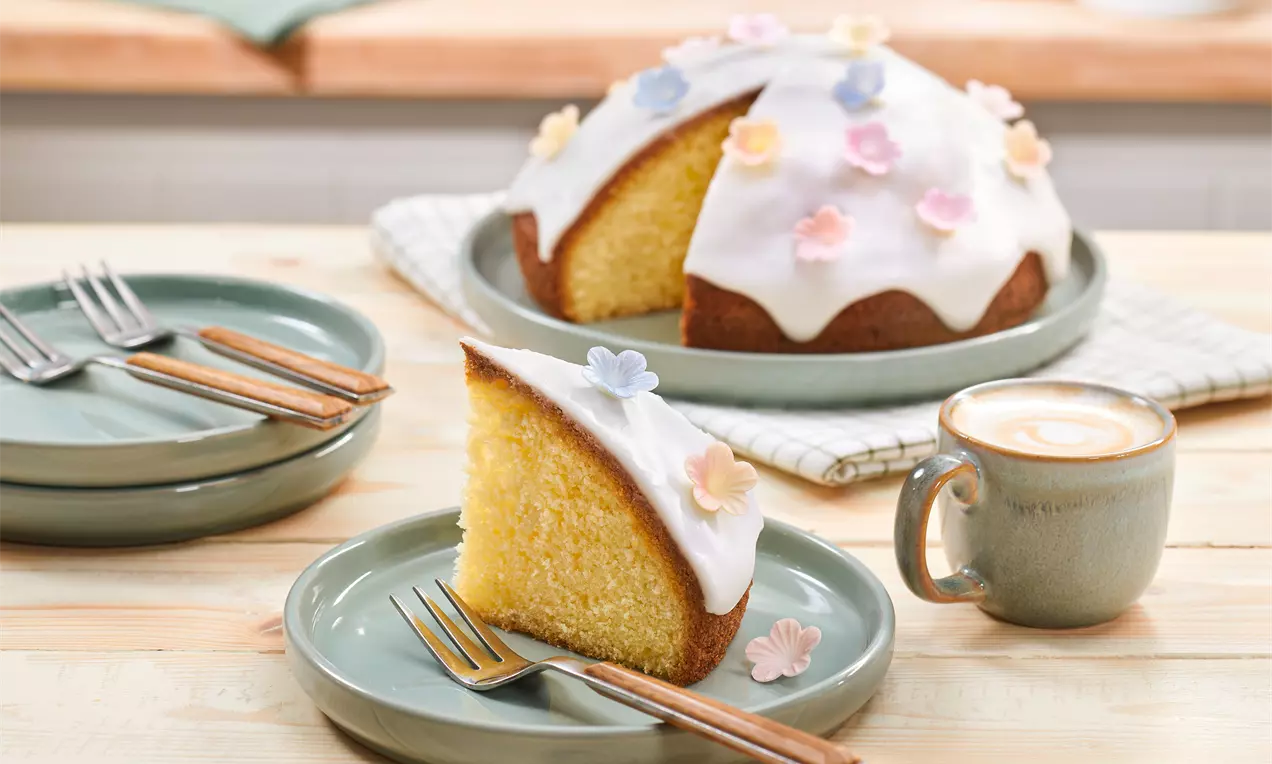 Saftiger, goldgelber Zitronenkuchen mit weißer Glasur und bunten Zuckerblüten, auf grünen Tellern mit Cappuccino in grüner Tasse angerichtet