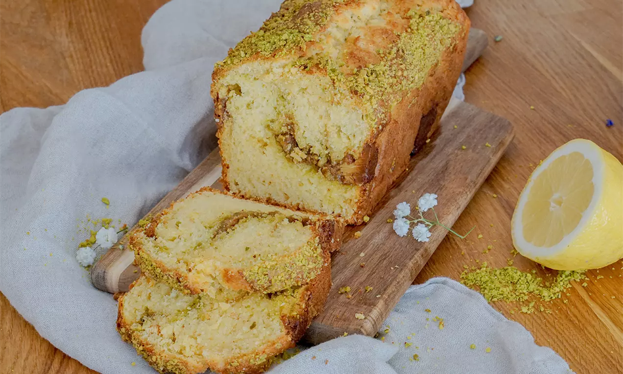 Ein länglicher Zitronen-Pistazienkuchen mit goldbrauner Kruste und grünem Pistazienstreusel, auf Holzbrett mit halber Zitrone und Stofftuch drapiert
