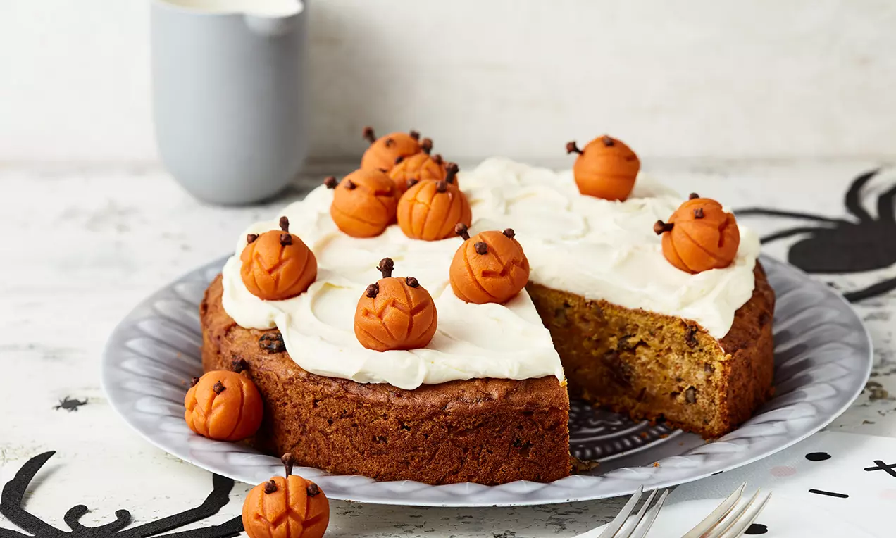 Saftiger Kürbiskuchen mit weißem Frosting, dekoriert mit kleinen Kürbissen aus orangefarbenem Fondant und Nelken auf grauer Platte