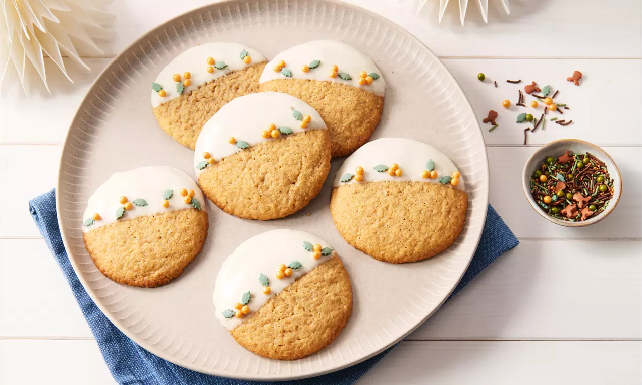 Runde Lebkuchen-Cookies mit weißer Glasur und bunter Zuckerdekoration auf beigefarbenem Teller, angerichtet auf blauer Stoffserviette