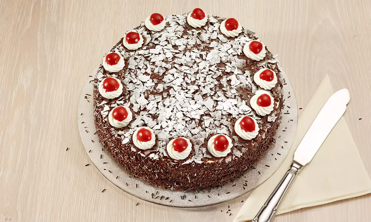 Torta rotonda ricoperta di scaglie di cioccolato e decorata con ciuffi di panna sormontati da ciliegie rosse su un piatto argentato