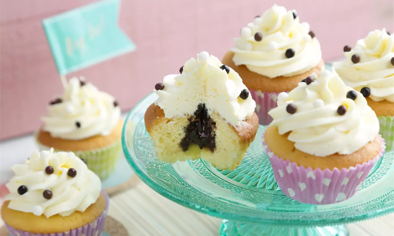 Vanillefarbene Cupcakes mit weißem Cream-Cheese-Frosting, Schokoladenkern und bunten Papierförmchen auf einer türkisfarbenen Glasplatte