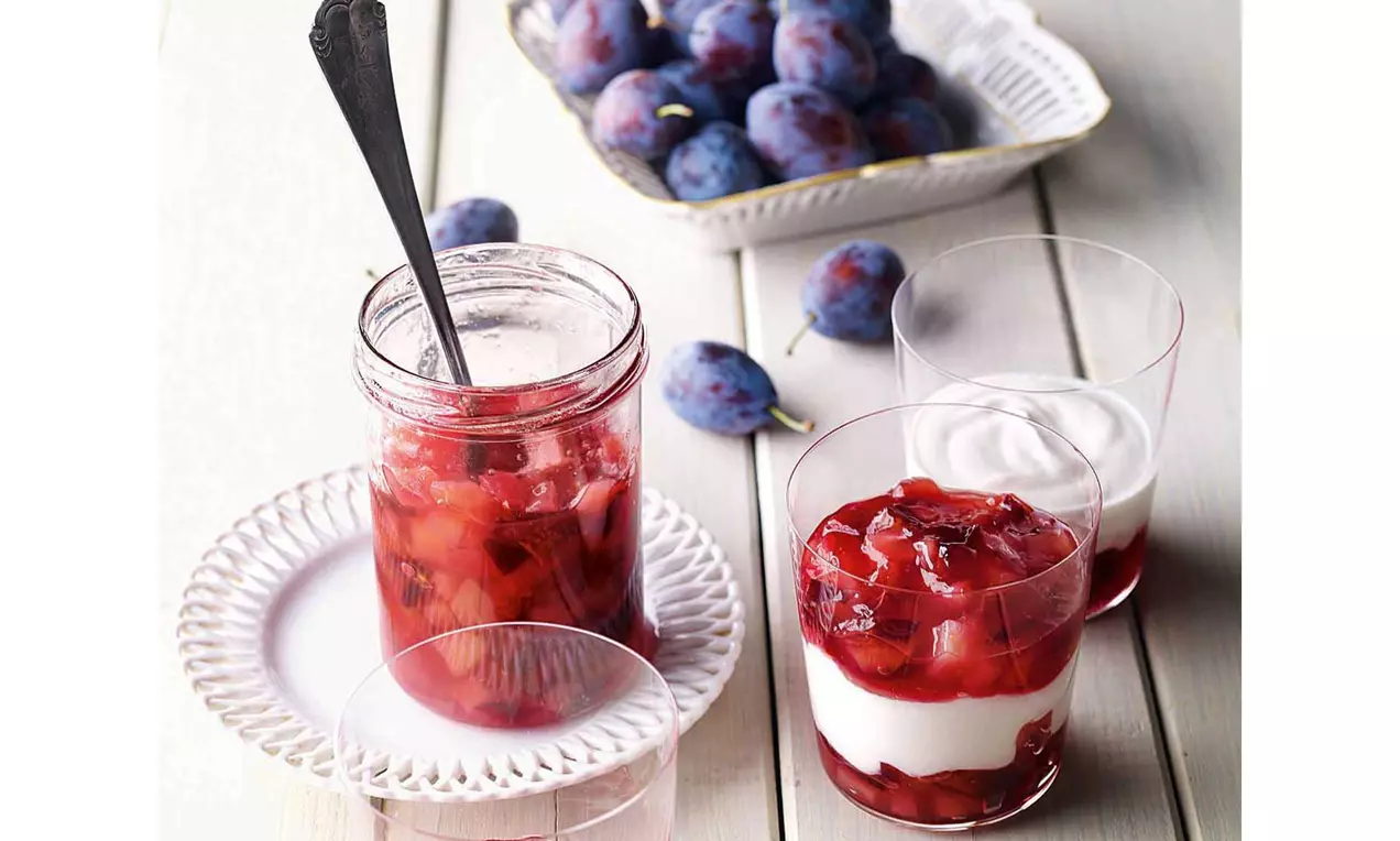 Composta rosso vivo di pere e prugne con pezzi morbidi, servita in un barattolo di vetro accanto a bicchieri con panna bianca su fondo chiaro