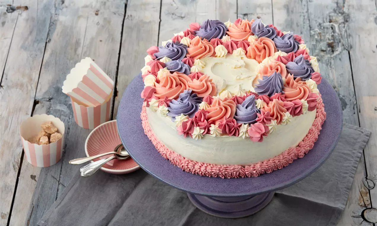 Runde Torte mit weißer Creme, umrandet von pastellfarbenen Blüten aus Rosa und Lila auf violetter Platte mit Holzuntergrund