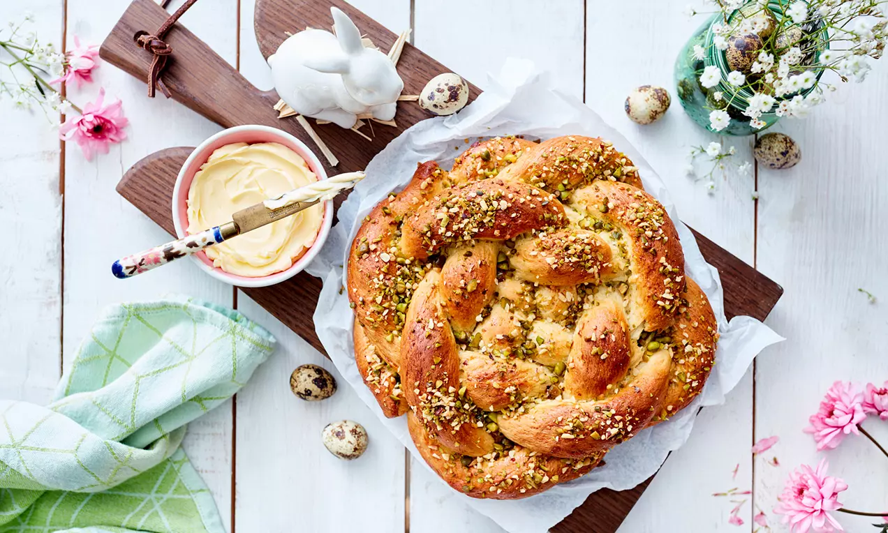 Goldbraune, geflochtene Knoten-Brötchen mit groben Pistazienstücken auf Butterpapier, angerichtet auf einem Holzbrett neben Butter und Frühlingsdeko
