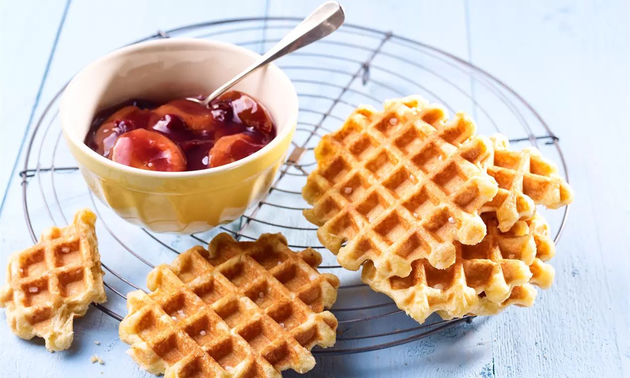 Goldgelbe Waffeln mit leichter Textur auf einem Metallrost neben einer cremefarbenen Schale mit glänzendem Pflaumen-Sauerkirsch-Kompott