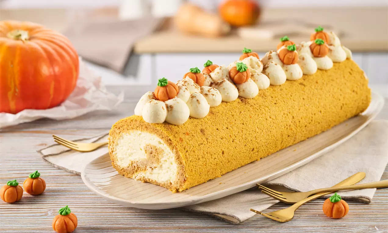 Eine goldbraune Biskuitrolle mit weißer Cremefüllung, dekoriert mit Sahnetupfen, Zimt und kleinen Zuckerkürbissen auf einer Platte.