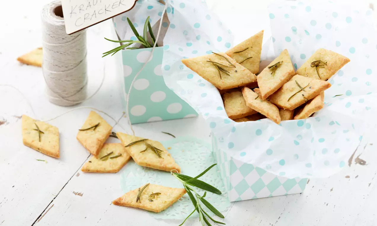 Knusprige Kräuter-Cracker in Rautenform mit goldbrauner Oberfläche und Rosmarinzweigen, präsentiert in einer türkis gemusterten Schachtel mit weißem Seidenpapier