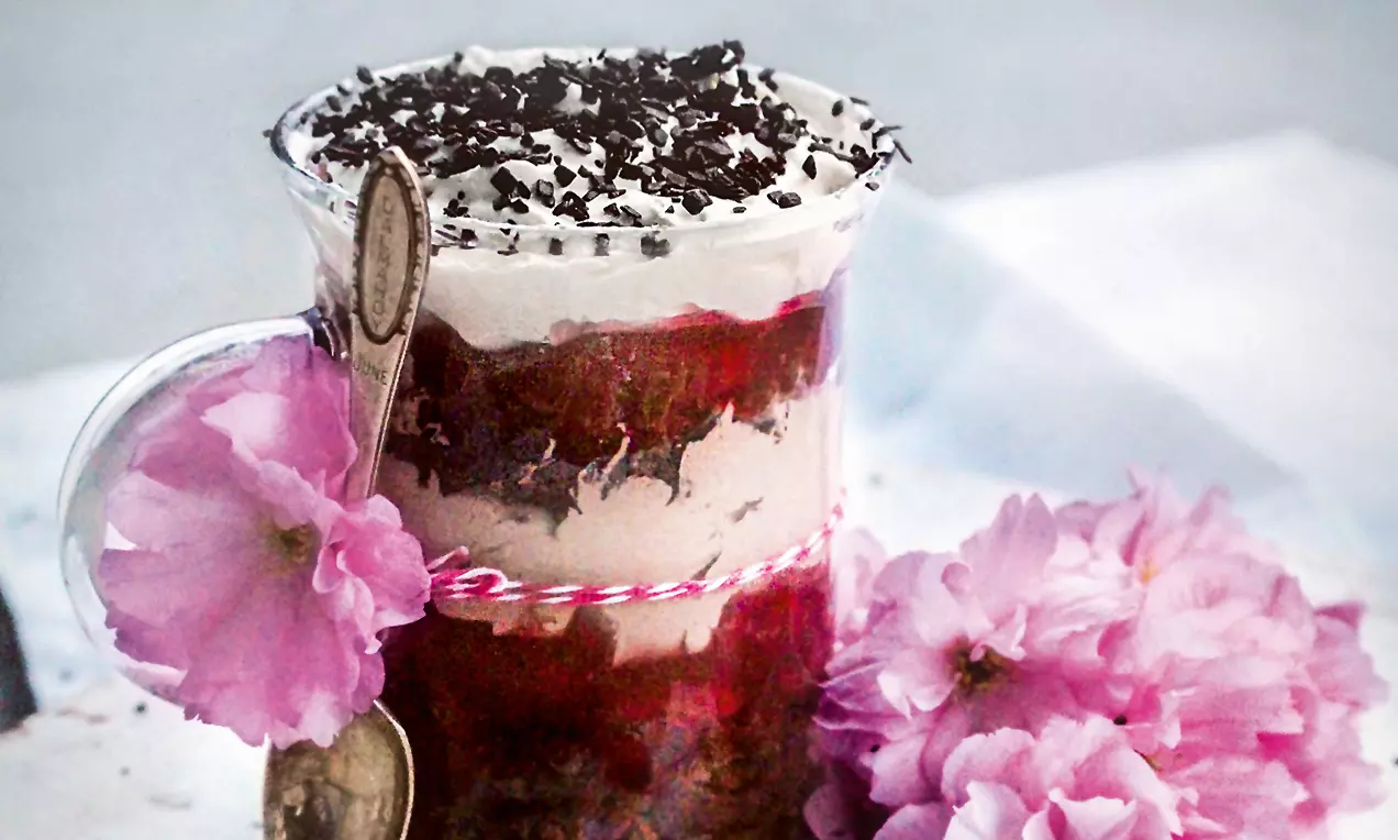 Dessert im Glas mit Schichten aus dunkler Creme, roter Fruchtmasse und Sahne, garniert mit Schokoraspeln, dekoriert mit rosa Blüten