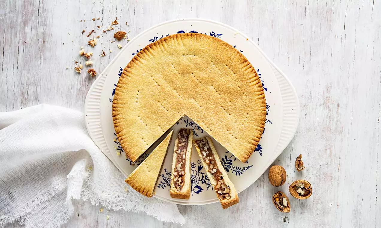 Crosta dorata dal bordo decorato racchiude un ripieno caramellato con pezzi di noce, disposta su un piatto bianco con motivi blu e noci accanto