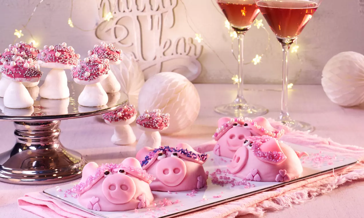 Zuckersüße Glücksschweine in Rosa mit feinen Details aus Zucker, dekoriert mit Glitzer und Perlen auf einem Teller neben Partydeko