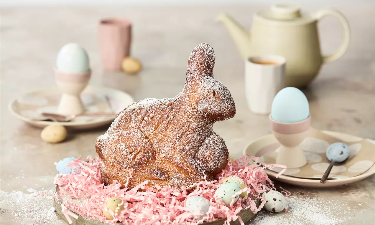 Ein brauner Osterhase aus Gebäck mit feinem Puderzucker bestäubt, auf rosa Papiergras umgeben von bunten Eiern und Geschirrarrangement