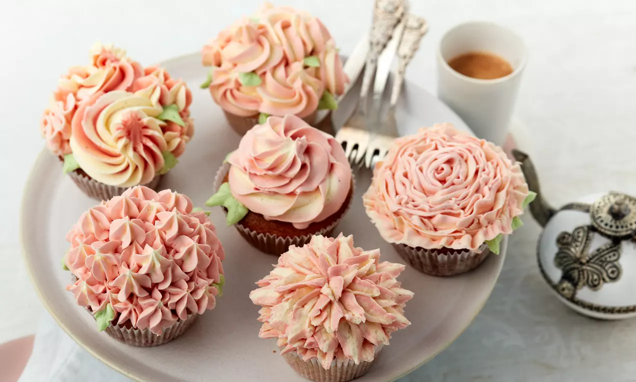 Cupcakes mit kunstvollen Blüten aus rosafarbenem, cremeweißem und zartgrünem Frosting auf einem flachen Teller mit Kaffee und Besteck