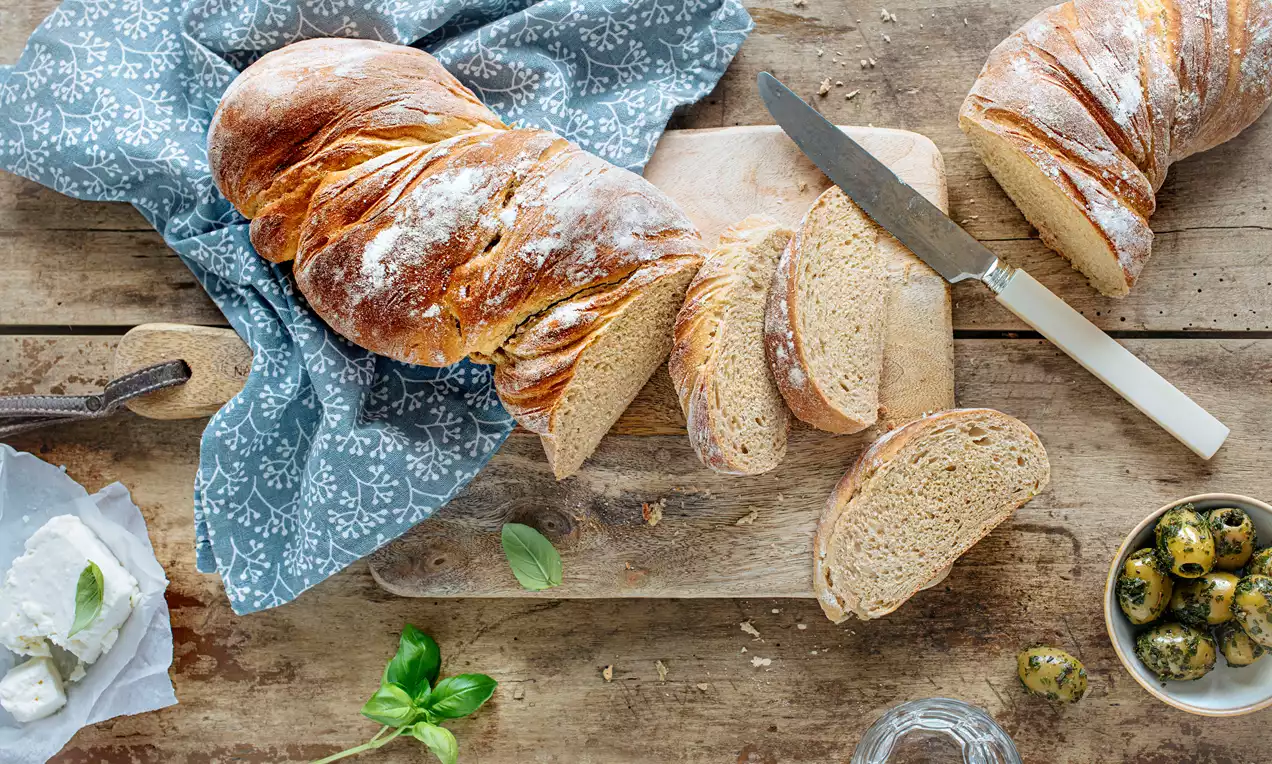 Rustikales Wurzelbrot aus Weizen und Roggen mit knuspriger Kruste und weicher Krume, teils aufgeschnitten auf Holzbrett mit Messer, Oliven und Kräutern