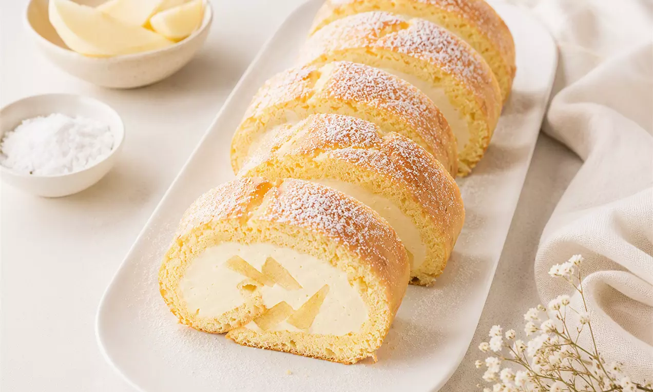 Goldgelbe Apfelroulade in Scheiben geschnitten, gefüllt mit heller Creme und Apfelstücken, bestäubt mit Puderzucker auf weißer Platte drapiert, AI-labeled image