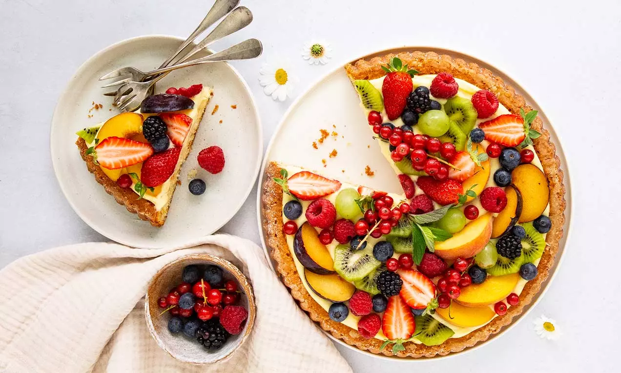 Früchtetorte mit goldbraunem Mürbeboden, hellem Belag und bunter Obstgarnitur aus Beeren, Trauben, Kiwi und Pfirsich auf weißem Teller